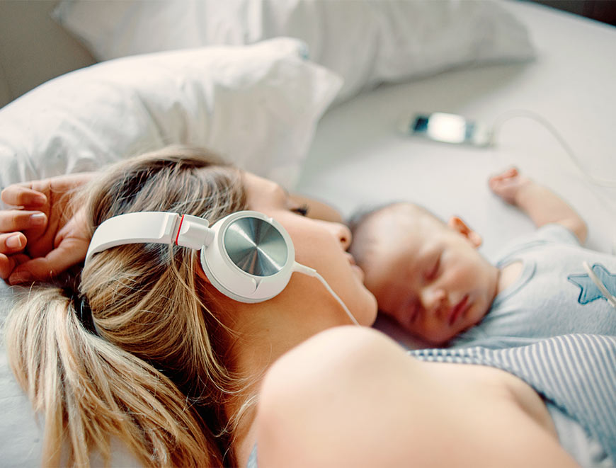 Photo of a mother wearing headphones laying beside a sleeping baby